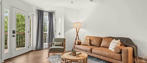 Una sala de estar con ventilador de techo, un sofá de cuero marrón, una mesa de centro de madera y un ventanal con cortinas.