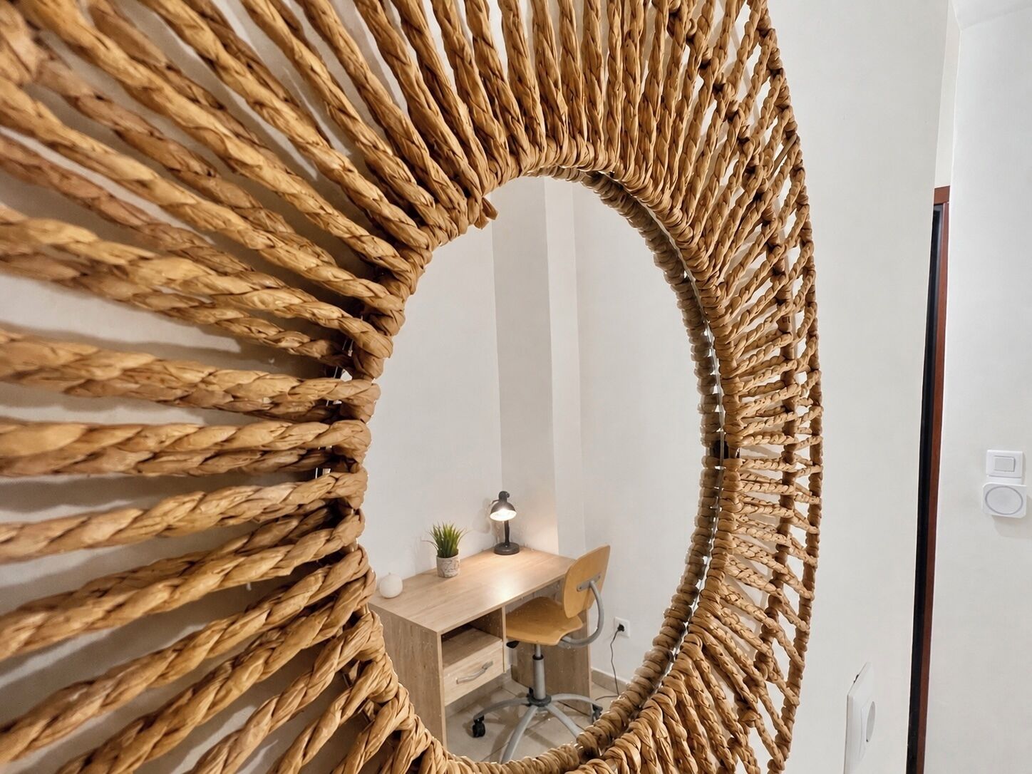 Une pièce avec un grand miroir à motifs tissés accroché au mur, un bureau en bois, une chaise et une petite plante.