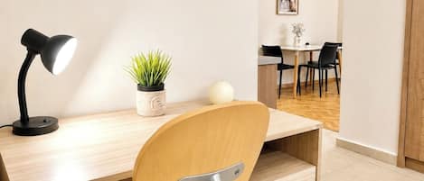 Un espace de bureau moderne comprenant un bureau, une chaise, une lampe, une plante en pot et, au fond, un coin repas.