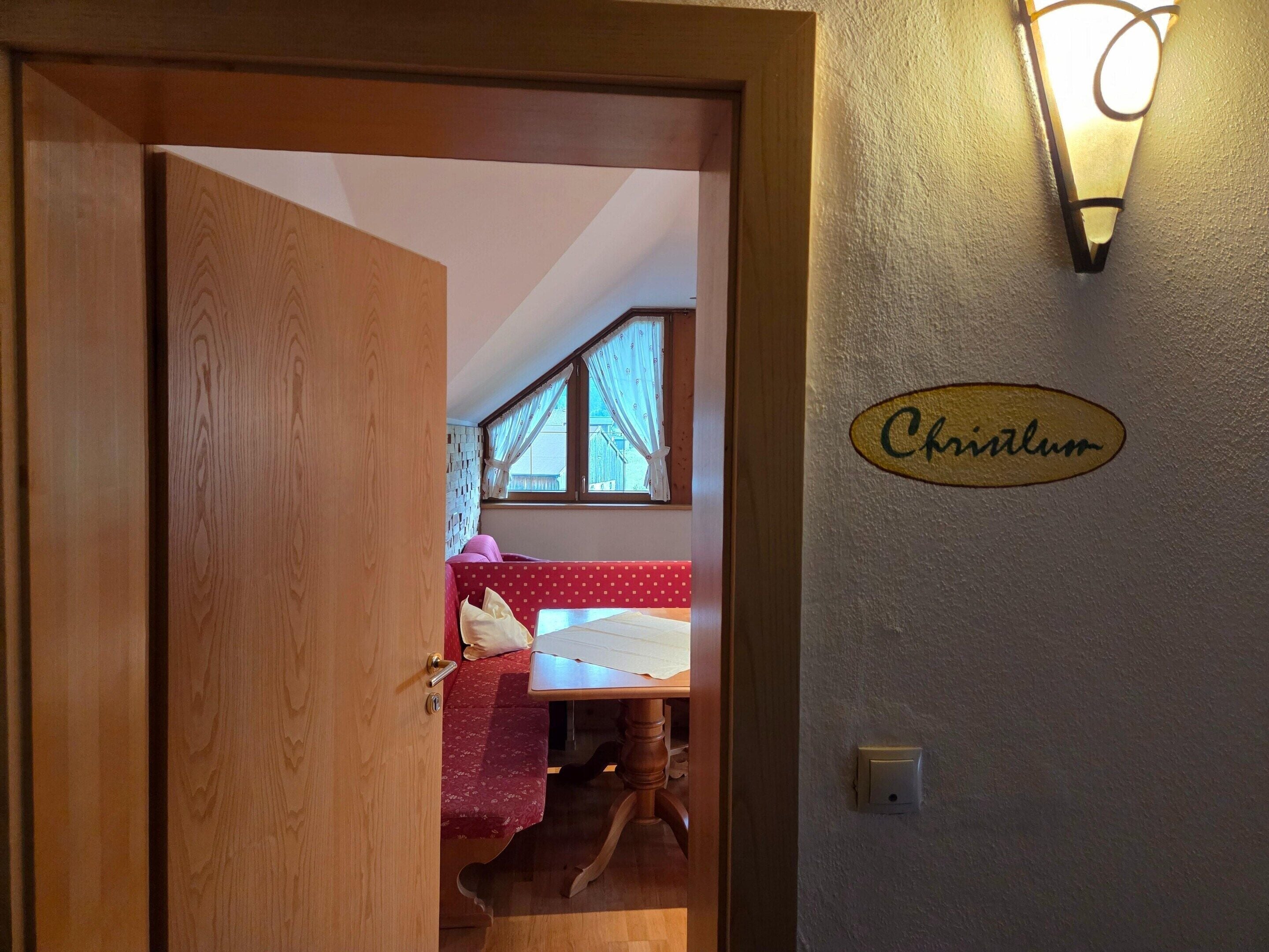 Ein Zimmer mit Holztür, einem Bett mit rot-weiß gemustertem Überwurf, einem kleinen Tisch und einer Wandleuchte mit dem Schriftzug 'Chritlum' an der Wand.