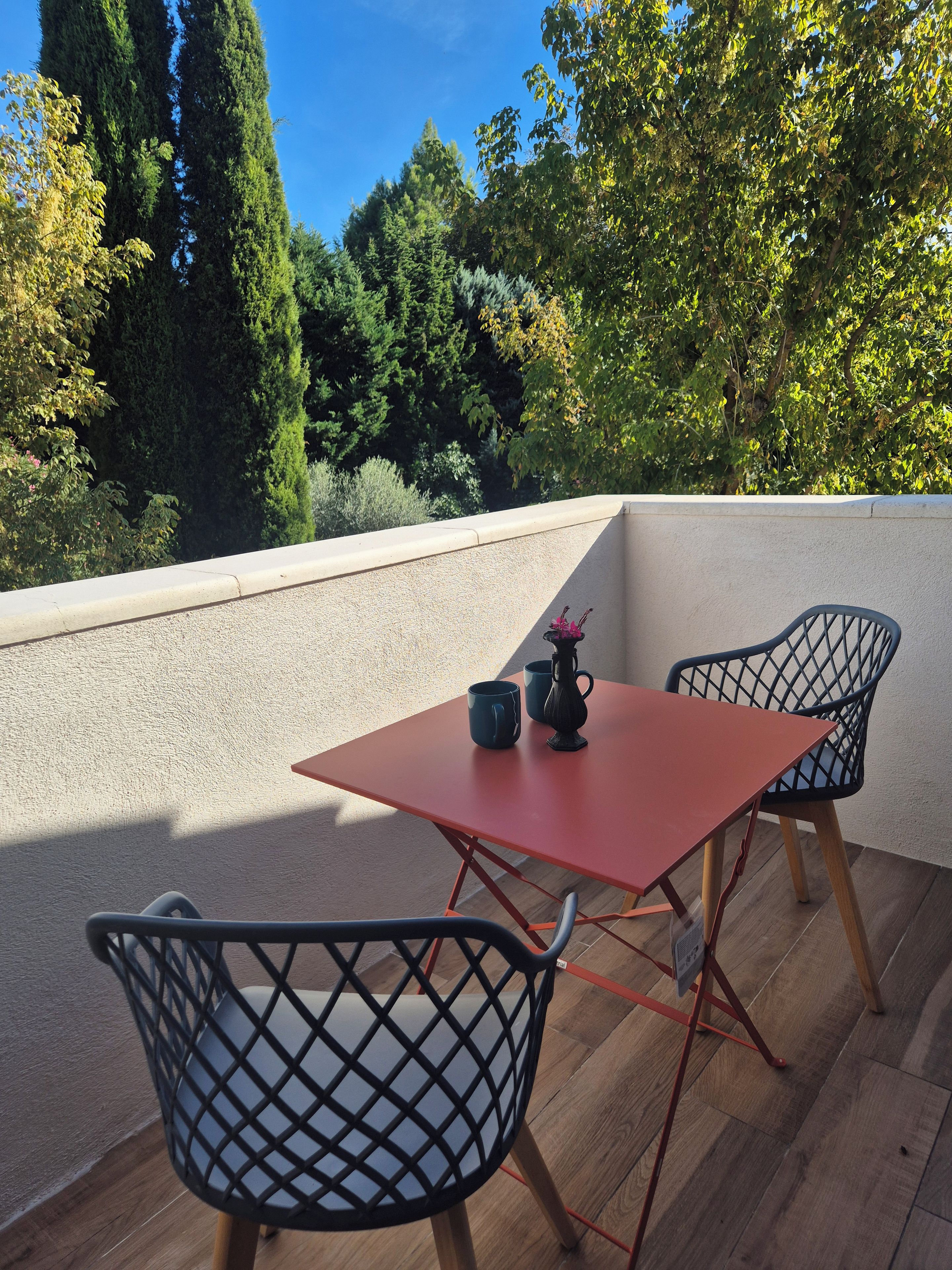 Un balcon avec une table rouge et deux chaises, donnant sur un jardin arboré et un ciel dégagé.