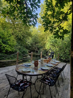 An outdoor dining area with a table set for four, surrounded by chairs, overlooking a forested area with a rope bridge.