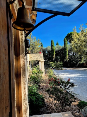 A bell on a wooden structure, a stone wall, and a garden with various plants and a pathway.