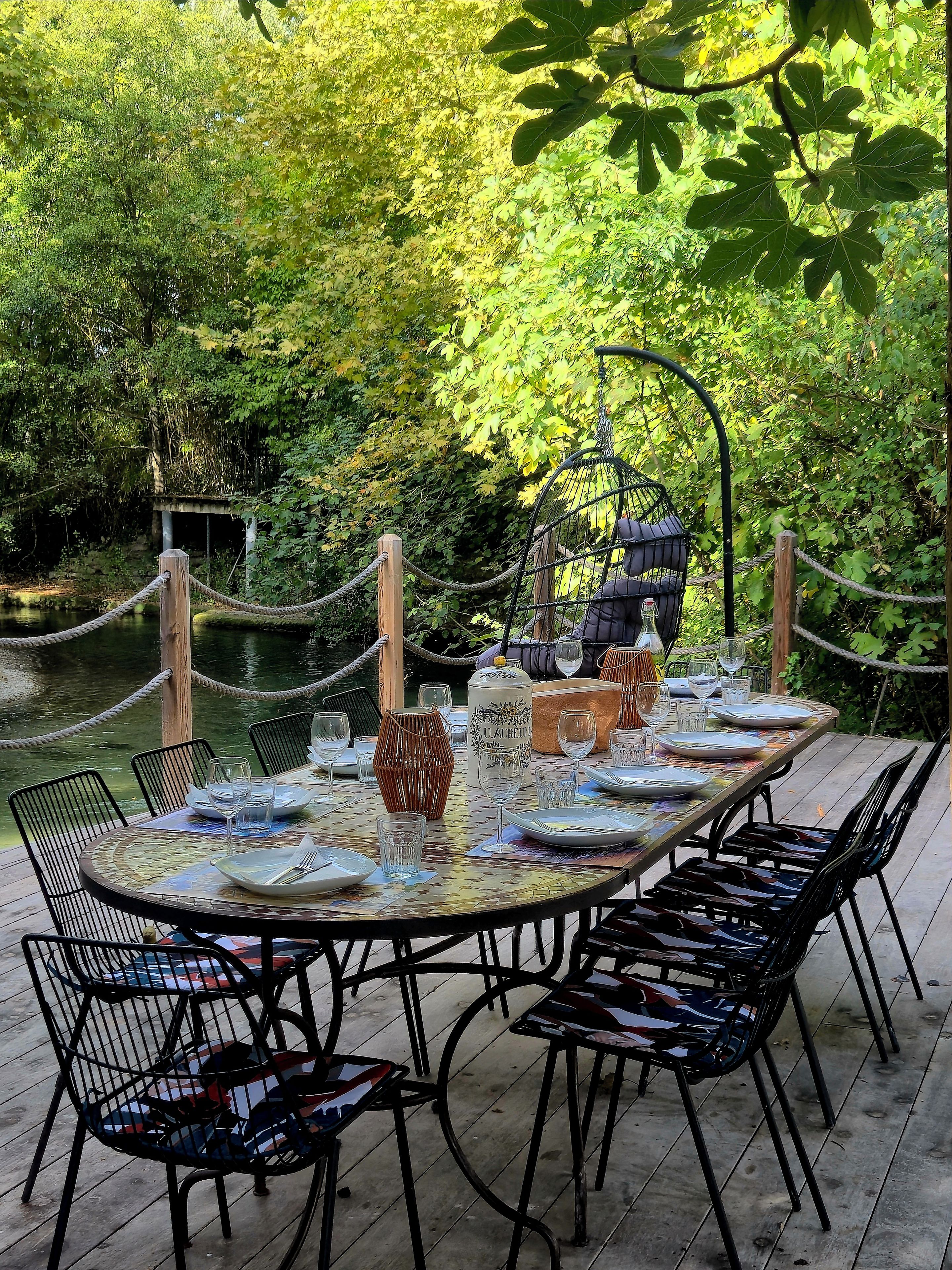 Un área de comedor con una mesa redonda preparada para seis personas, rodeada de sillas, una barrera de cuerda y vista a un río.