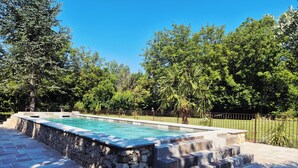 A rectangular swimming pool with stone steps leading up to it, surrounded by a stone wall and a metal fence.