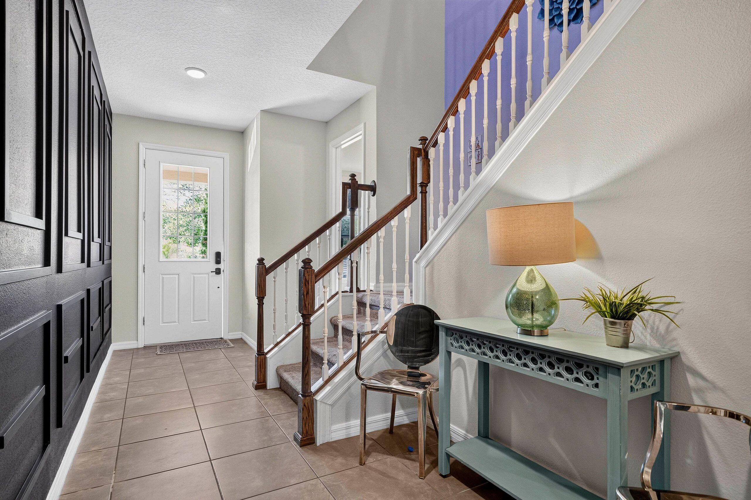 A hallway with a wooden staircase, a door with a window, and a console table with a lamp and a plant.
