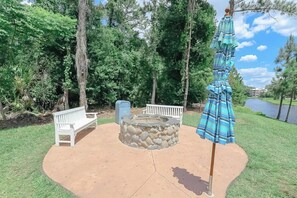 A patio area with a stone fire pit, a white bench, and a blue-striped umbrella. - Orlando | Laid-Back Island Escape | Pool + Golf (Lake Buena Vista)