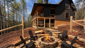 Eine Holzhütte mit einem Steinkamin und Adirondack-Stühlen, umgeben von einer Holzterrasse und einem Wald.