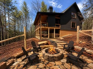 Eine Holzhütte mit einem Steinkamin und Adirondack-Stühlen, umgeben von einer Holzterrasse und einem Wald.