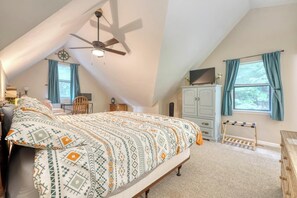 Quarto com cama, ventoinha no teto, janela com cortinas, cômoda e uma pequena mesa.
