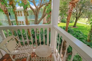 A porch with a white railing, a bench, and a table. In the background, there are buildings with green and white facades and trees. - Orlando | Relaxed Island Family Stay | Pool + Golf | 2BR (Lake Buena Vista)