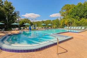 A swimming pool with a tiled edge, surrounded by lounge chairs and umbrellas, with trees in the background. - Orlando | Relaxed Island Family Stay | Pool + Golf | 2BR (Lake Buena Vista)