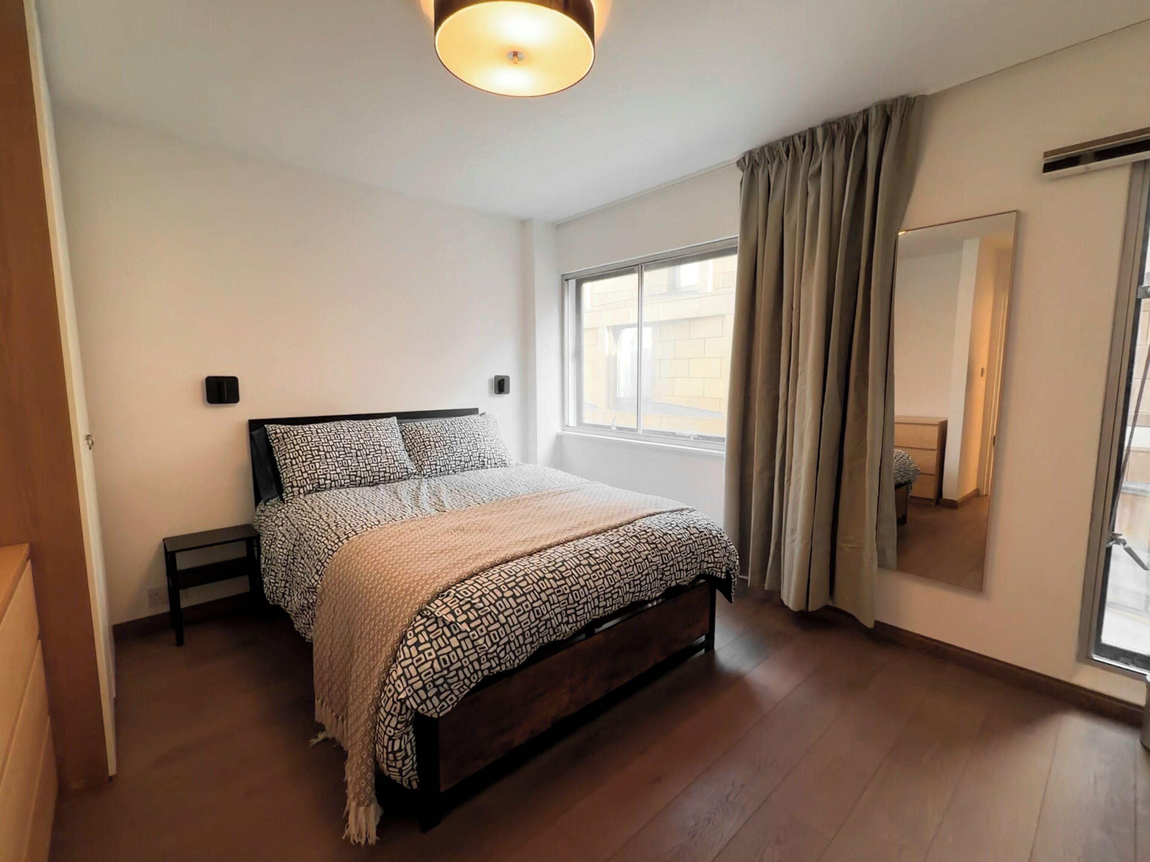Un dormitorio con cama, mesita de noche, ventana con cortinas y espejo en la pared.