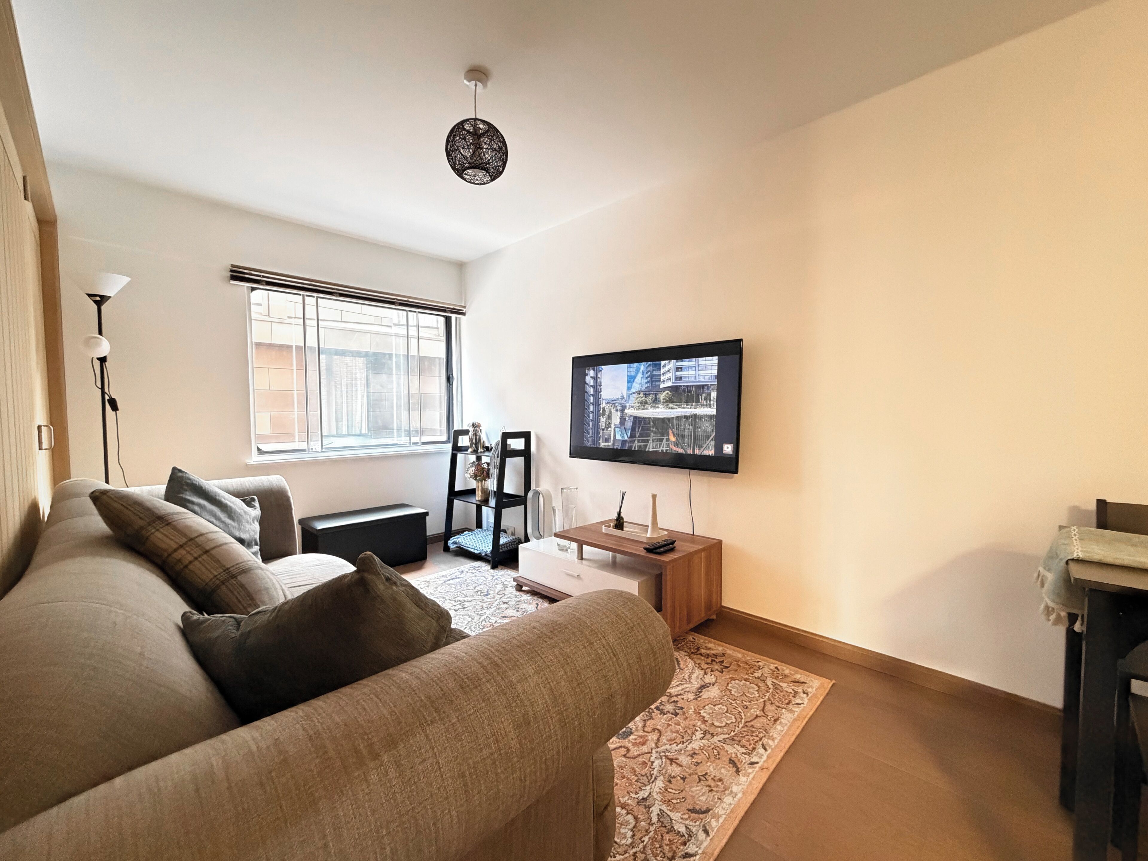 A modern living room with a sofa, a flat-screen TV mounted on the wall, a floor lamp, and a window with blinds.