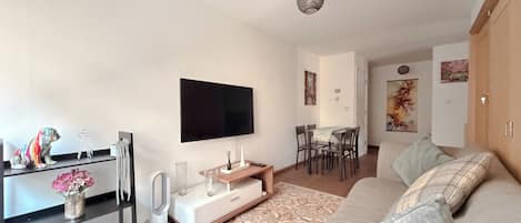 A modern living room with a flat-screen TV, a sofa, a dining table with chairs, and a decorative vase with flowers.