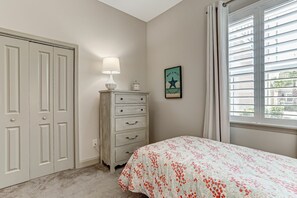 A bedroom with a bed, a dresser, a lamp, a framed quote, and a window with blinds. - Amelia Island Luxury Summerwoods Condo near Ritz Carlton / Omni (Fernandina Beach)
