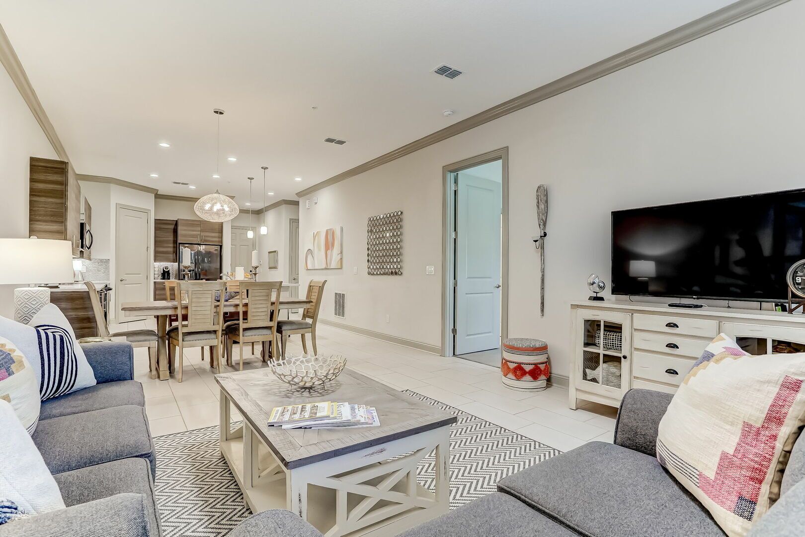 Een moderne woonkamer met een bank, salontafel en televisie. Er is een eethoek met stoelen en een tafel. De kamer heeft een neutrale kleurenpalet en beschikt over een grote spiegel en kunstwerken aan de wanden.