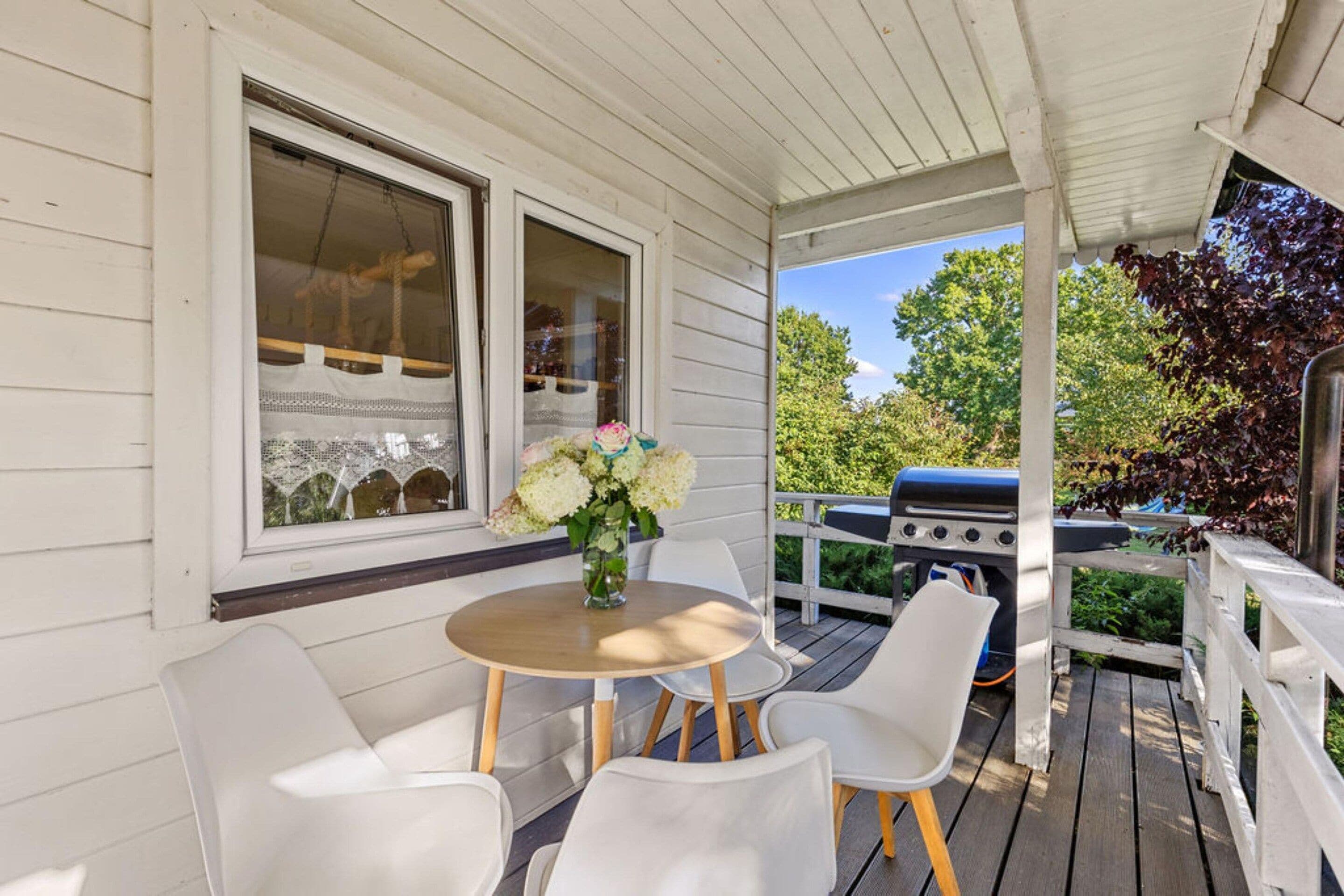 A covered porch with a table and chairs, a grill, and a window with a view of trees.