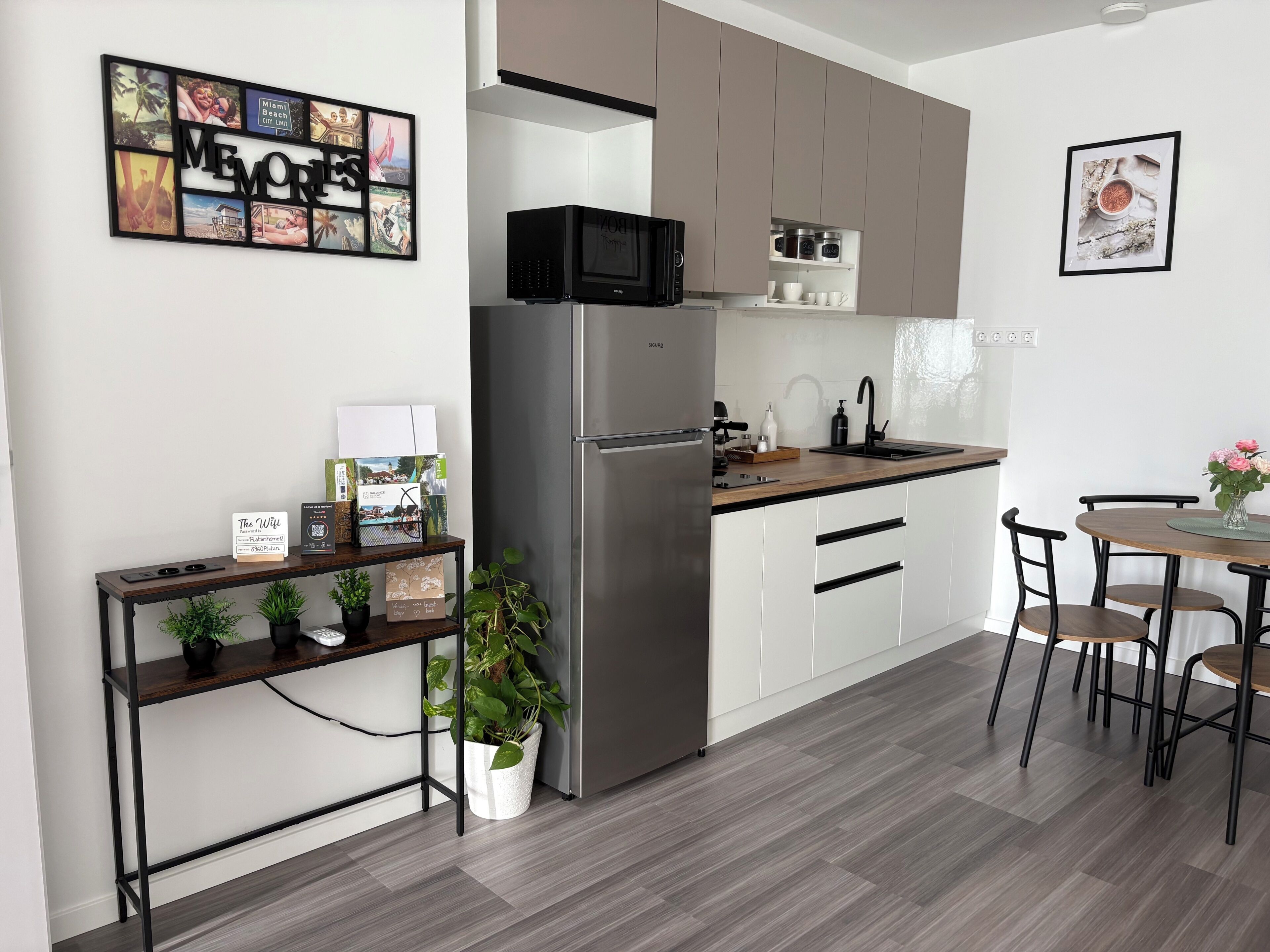 A modern kitchen with a stainless steel refrigerator, a microwave, and a sink. There is a dining table with chairs and a potted plant.