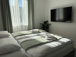 A hotel room with a bed, a TV mounted on the wall, and a window with sheer curtains.