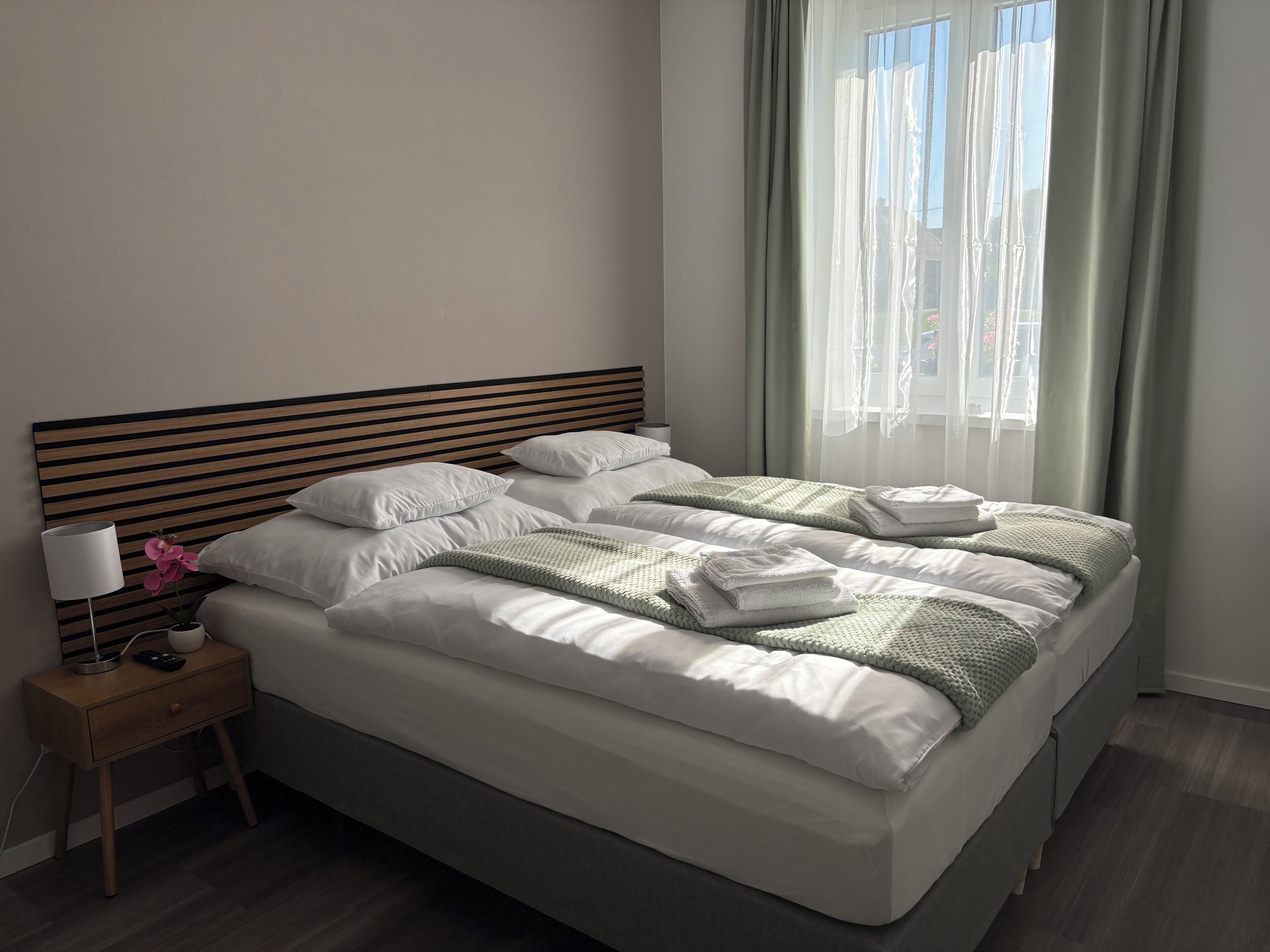 A neatly made bed with white linens and a green knitted blanket, a bedside lamp, and a small potted orchid on a wooden nightstand.