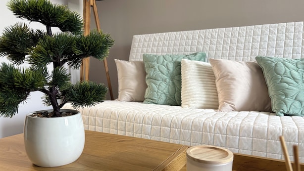 Une pièce chaleureuse avec un canapé blanc matelassé, un bonsaï en pot et une bougie parfumée posée sur une table en bois.