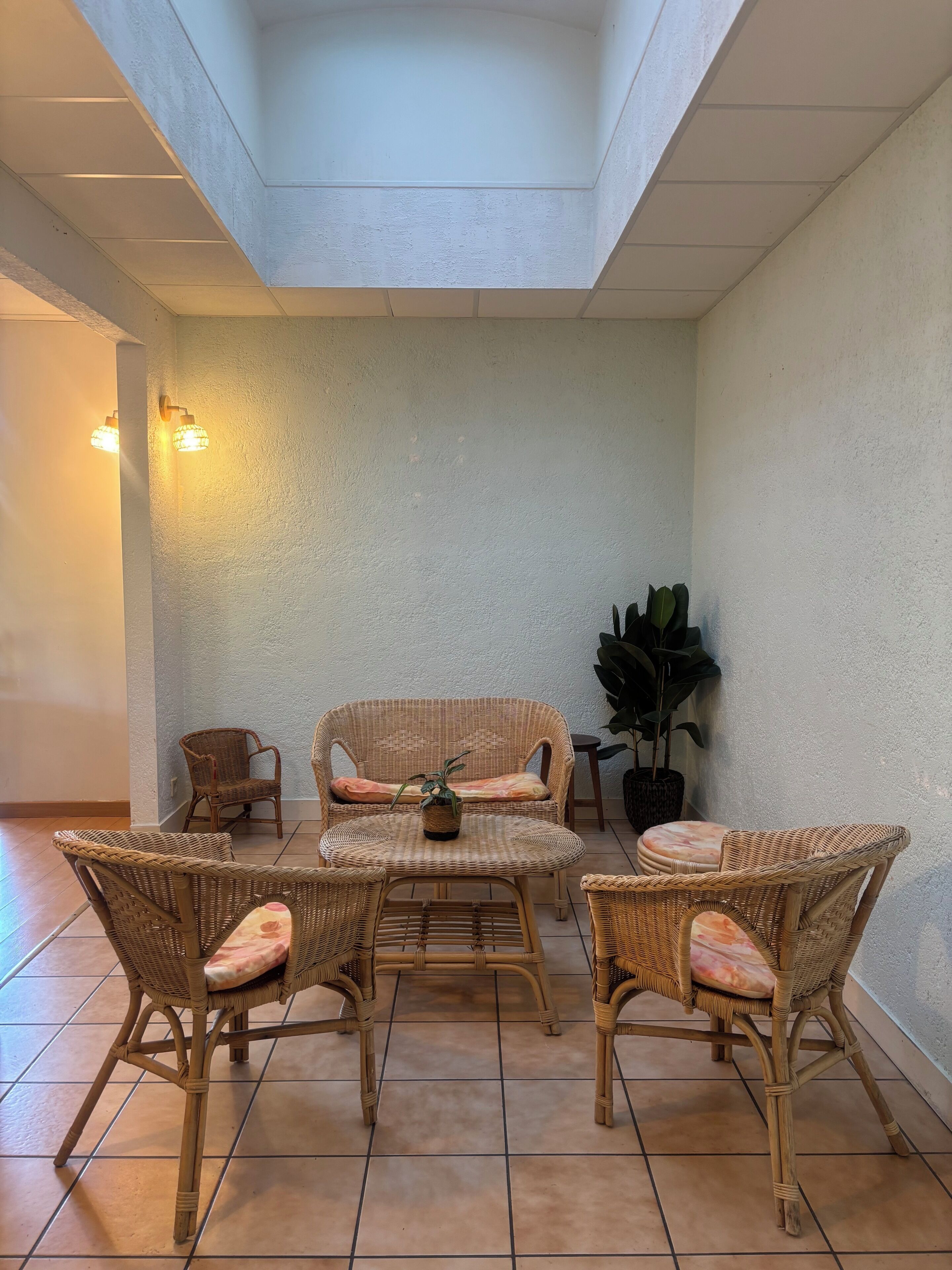 Una habitación con techo alto, piso de baldosas y una zona de estar con muebles de mimbre y una pequeña mesa.