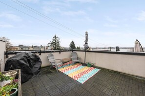 A rooftop terrace with a patterned rug, two chairs, a table, a grill, and a view of the cityscape.