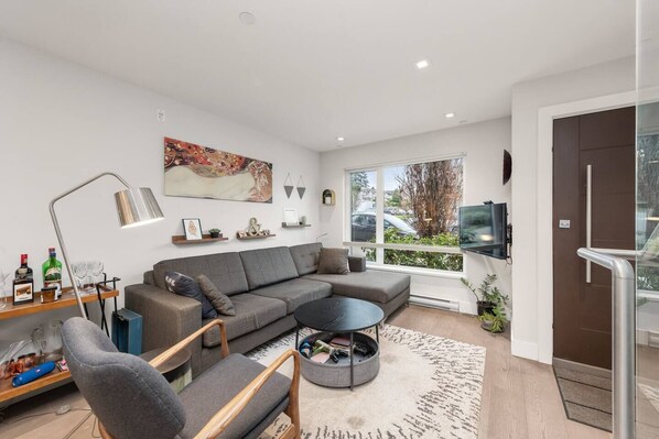 A modern living room with a grey sectional sofa, a round coffee table, and a TV mounted on the wall. There is a large window providing a view of the outside.