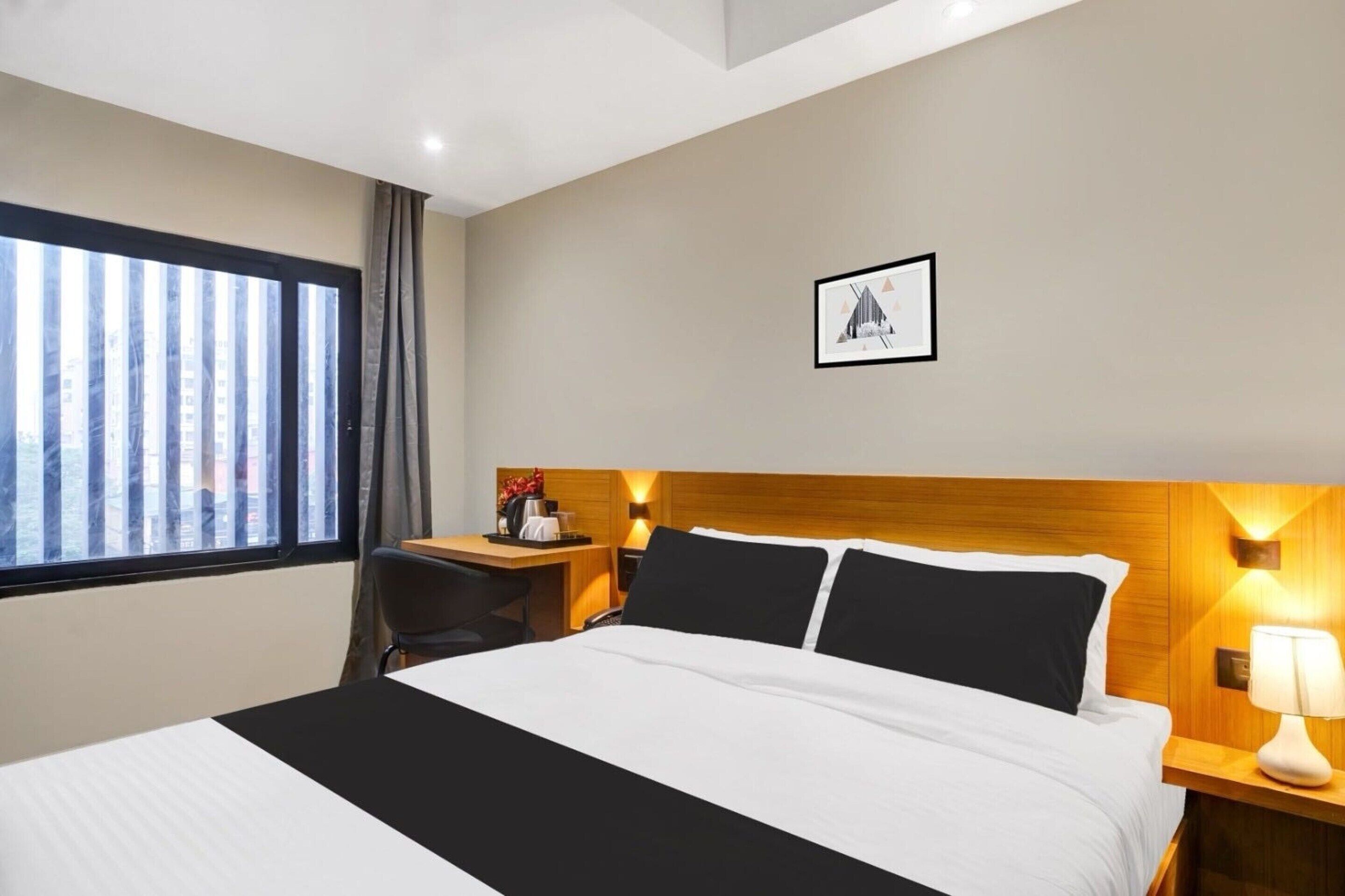 Habitación de hotel con cama, escritorio, silla, ventana con cortinas, un cuadro en la pared y mesita de noche con lámpara.