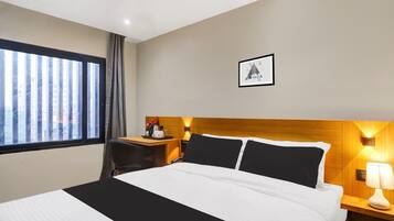 Habitación de hotel con cama, escritorio, silla, ventana con cortinas, un cuadro en la pared y mesita de noche con lámpara.