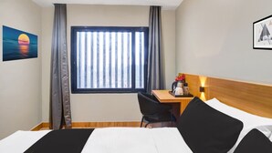 Hotellihuone, jossa on sänky, työpöytä, tuoli, pystysuorilla lamelleilla varustettu ikkuna ja taulu seinällä.