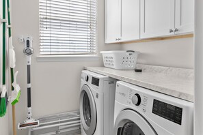 A laundry room with two front-loading washing machines, a drying rack, and a window with blinds. - Blue Chip Escape-Private Pool-30A-Bikes (Santa Rosa Beach)
