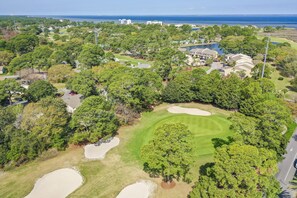 Luftaufnahme eines Golfplatzes mit Sandbunkern, Bäumen und Gebäuden im Hintergrund.