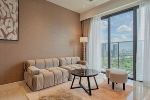 Una sala de estar moderna con un sofá color beige, una mesa de centro redonda y vistas de un paisaje urbano a través de ventanas grandes.