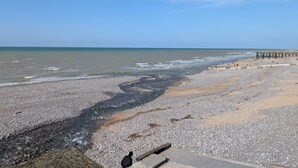 En stenet strand med en træbro, der strækker sig ud i vandet, klare himmel og roligt hav.