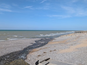 Ein Kiesstrand mit einem hölzernen Steg, der sich ins Wasser erstreckt, bewölkter Himmel und ruhige See.