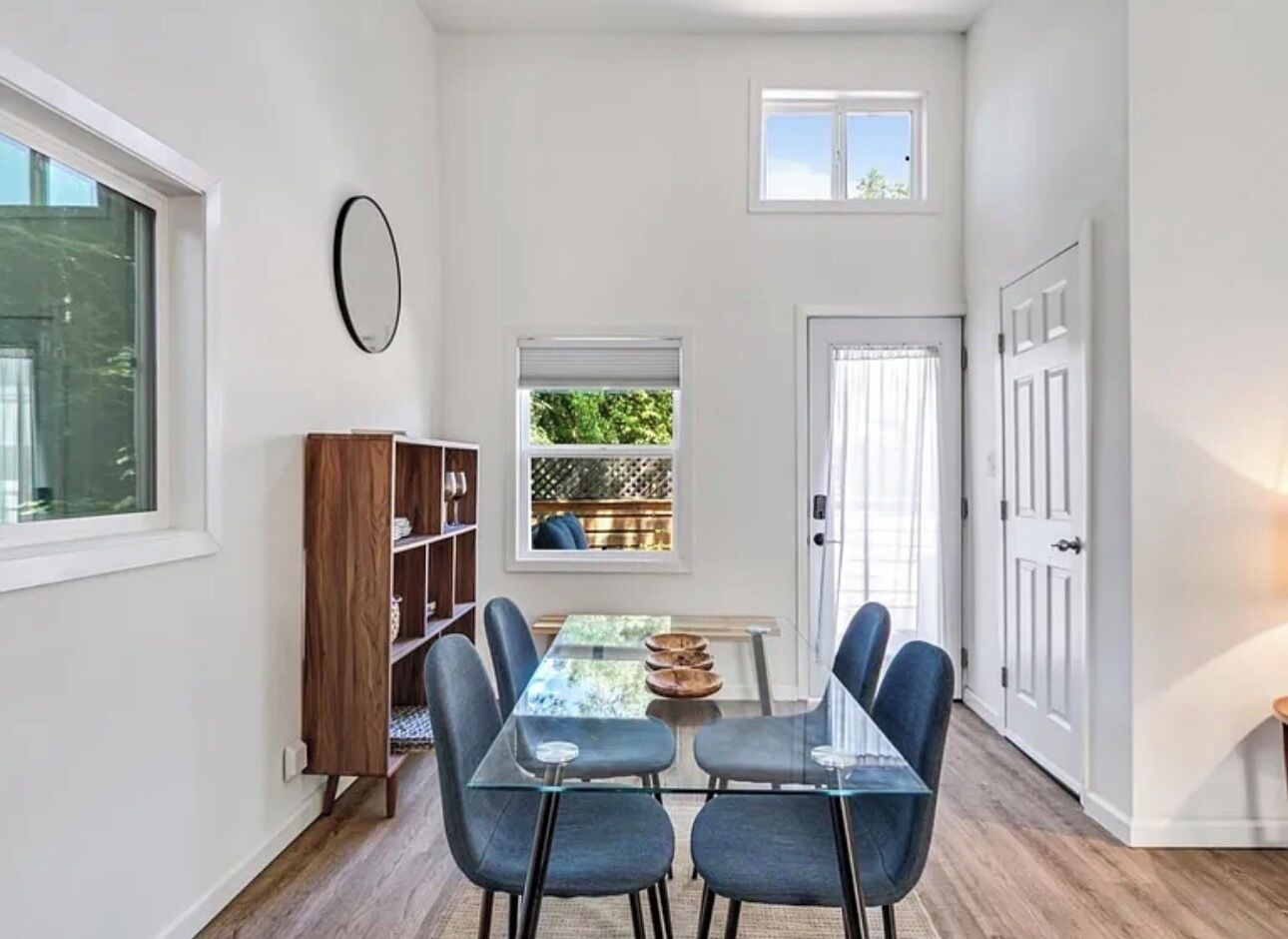 A dining area with a glass table and chairs, a wooden bookshelf, and a window with a view of greenery.