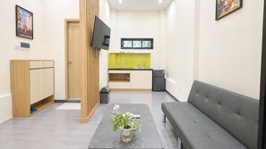 Uma sala de estar moderna com um sofá cinza, uma mesinha de centro com uma planta, televisão na parede e uma área de cozinha com painel verde.