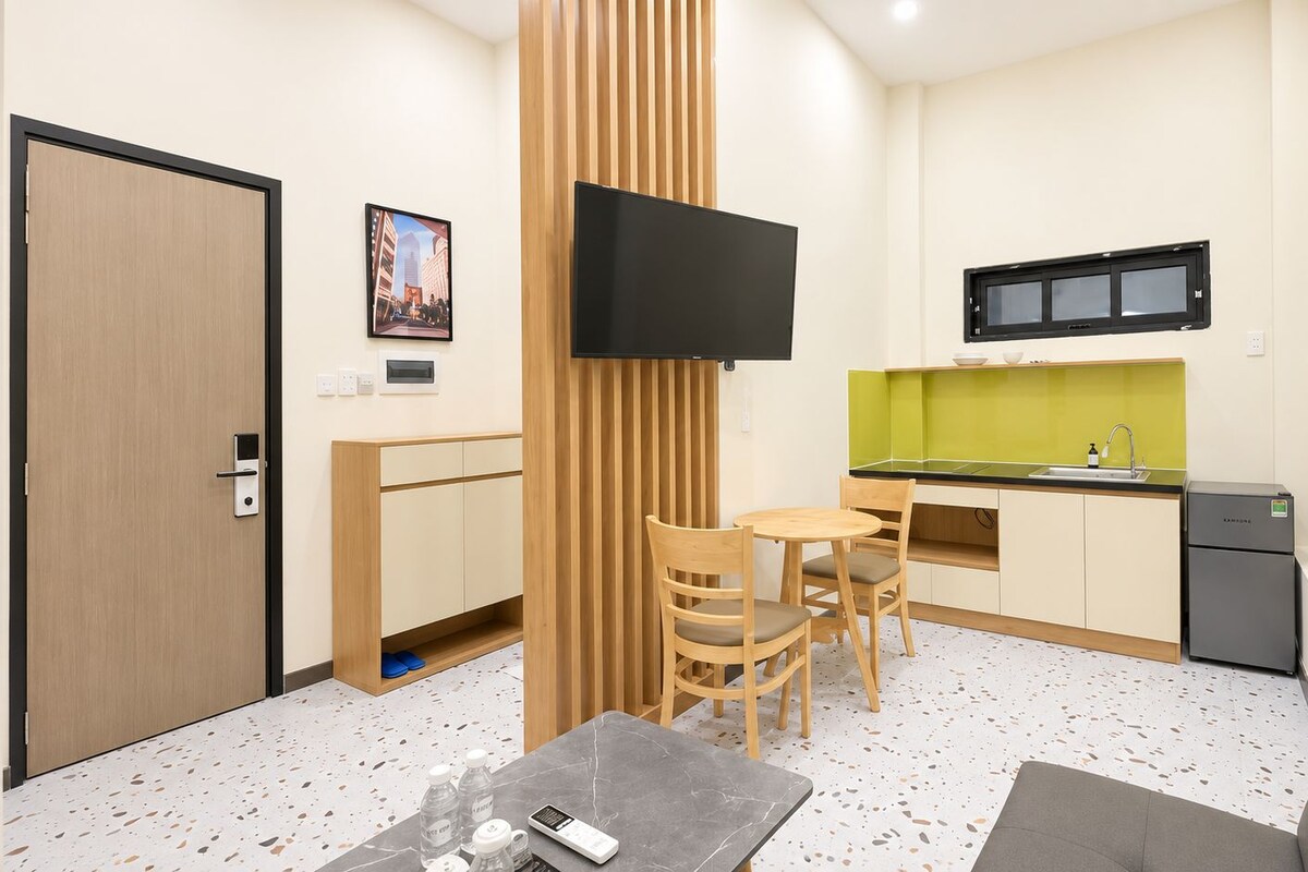 Quarto de hotel compacto com cozinha compacta, área de refeições com duas cadeiras, sofá, TV fixada na parede e porta com puxador.