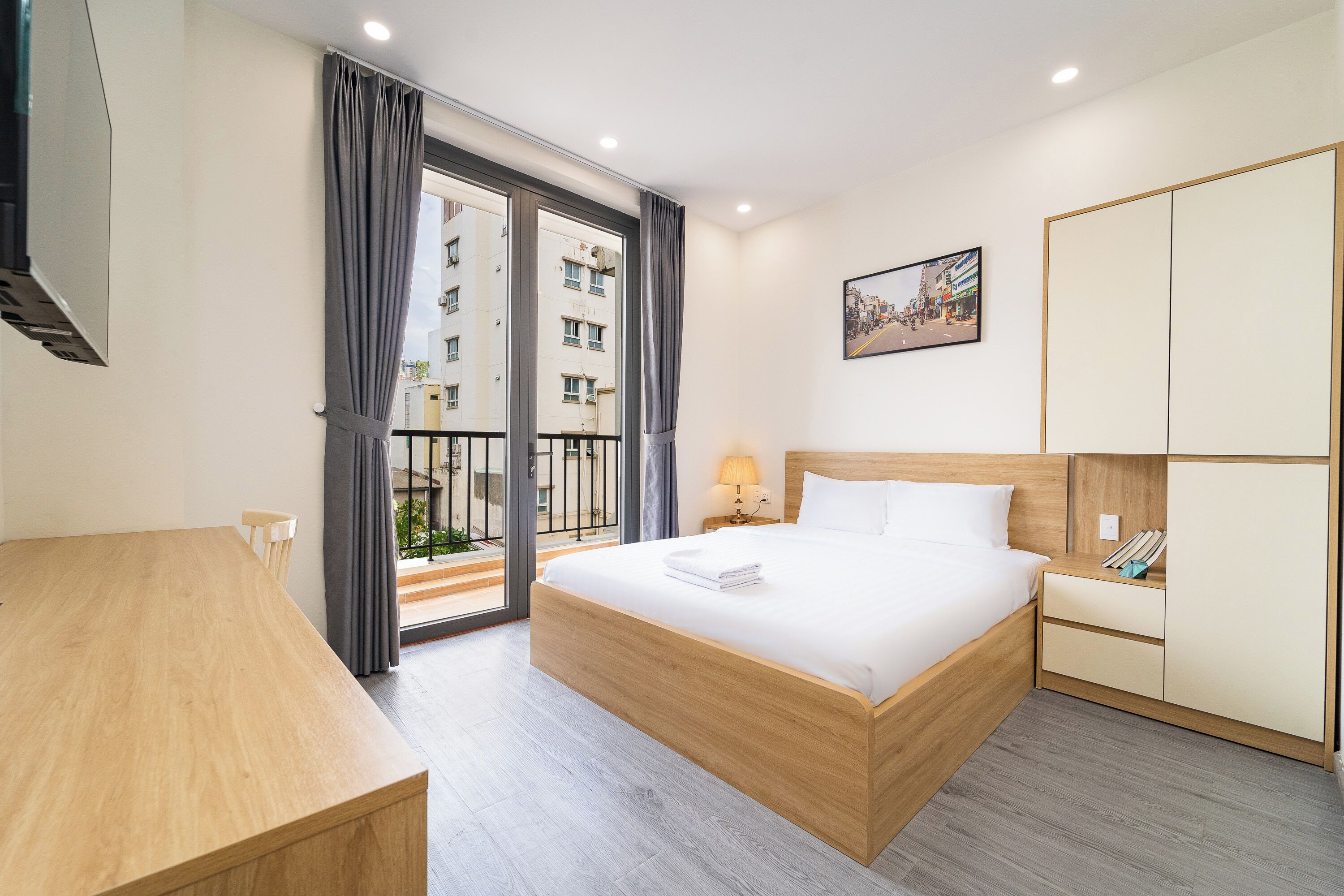 A hotel room with a bed, a desk, a nightstand, a lamp, a wardrobe, and a view of the outside.