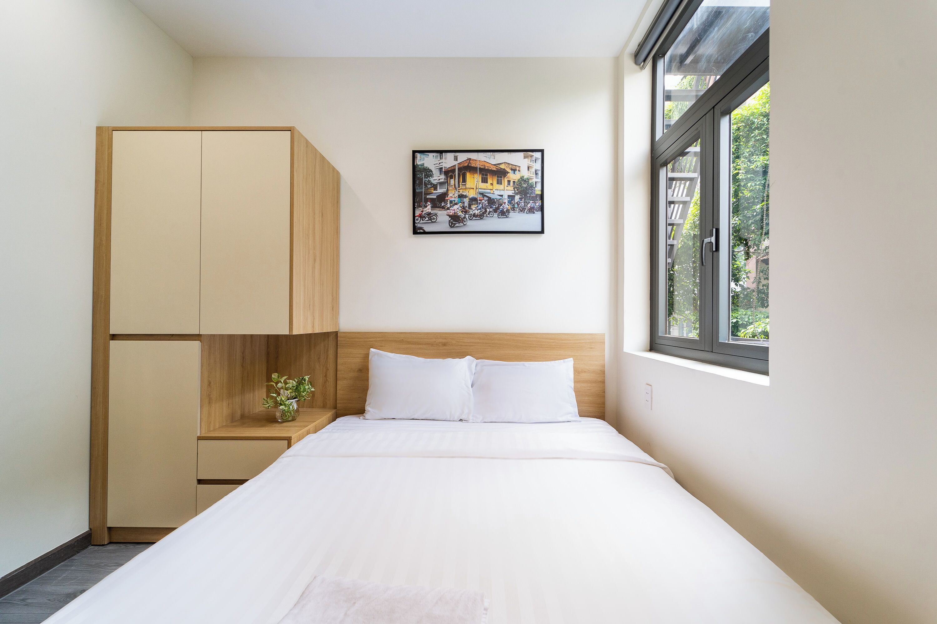 A bedroom with a bed, a wooden wardrobe, a small plant, and a window with a view of buildings.