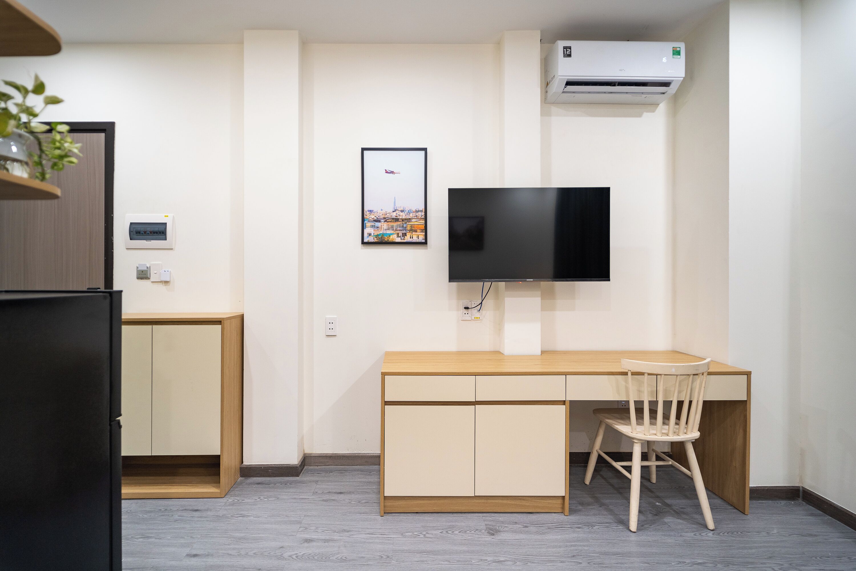 A compact room with a wooden desk, a chair, a television mounted on the wall, and an air conditioning unit.