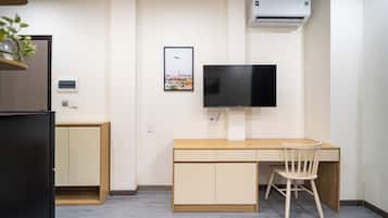 A compact room with a wooden desk, a chair, a television mounted on the wall, and an air conditioning unit.