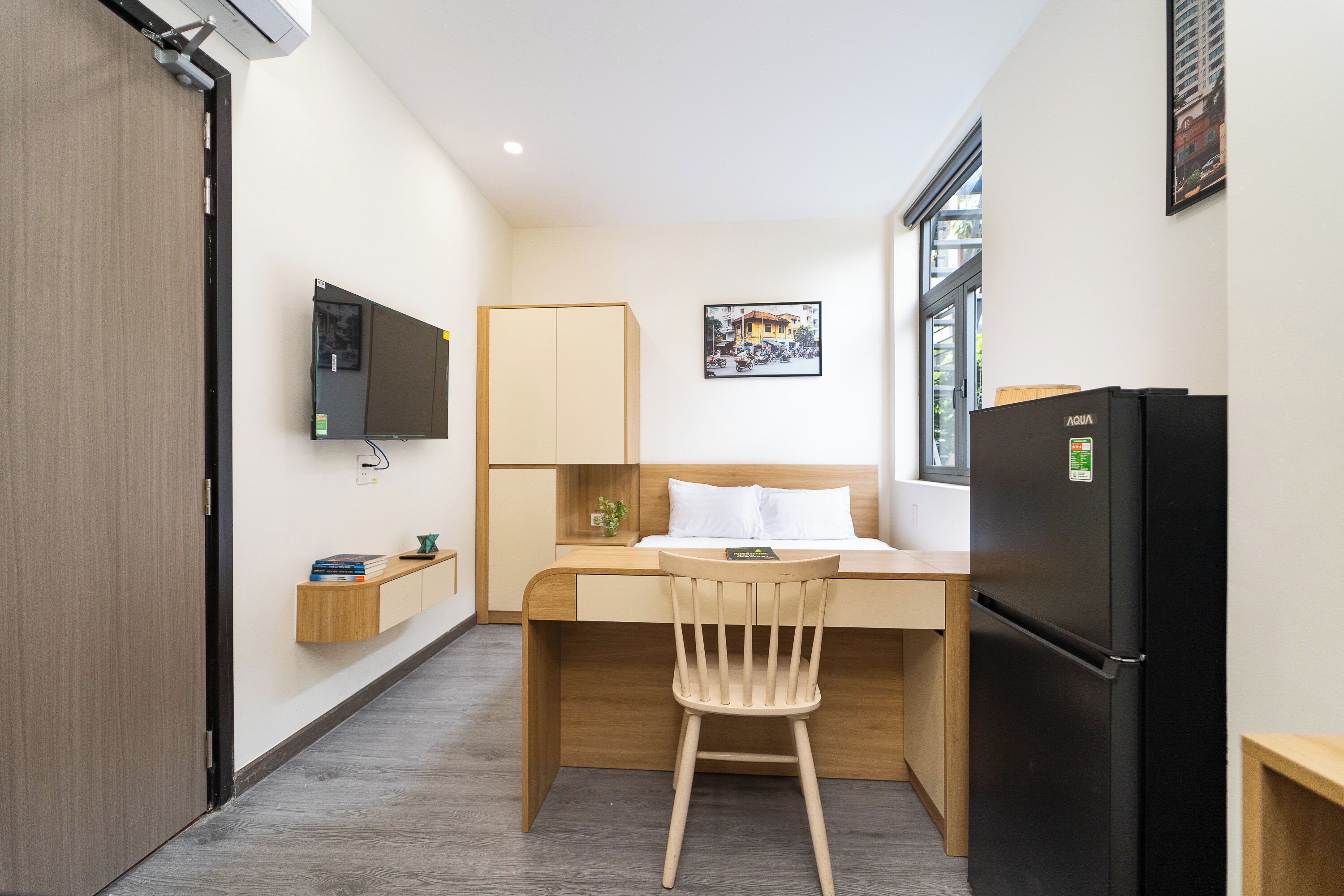 A compact hotel room with a bed, a desk, a chair, a TV, a refrigerator, and a window.