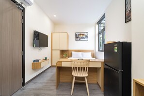 Quarto de hotel pequeno com cama, escrivaninha, cadeira, televisão, frigorífico e janela.