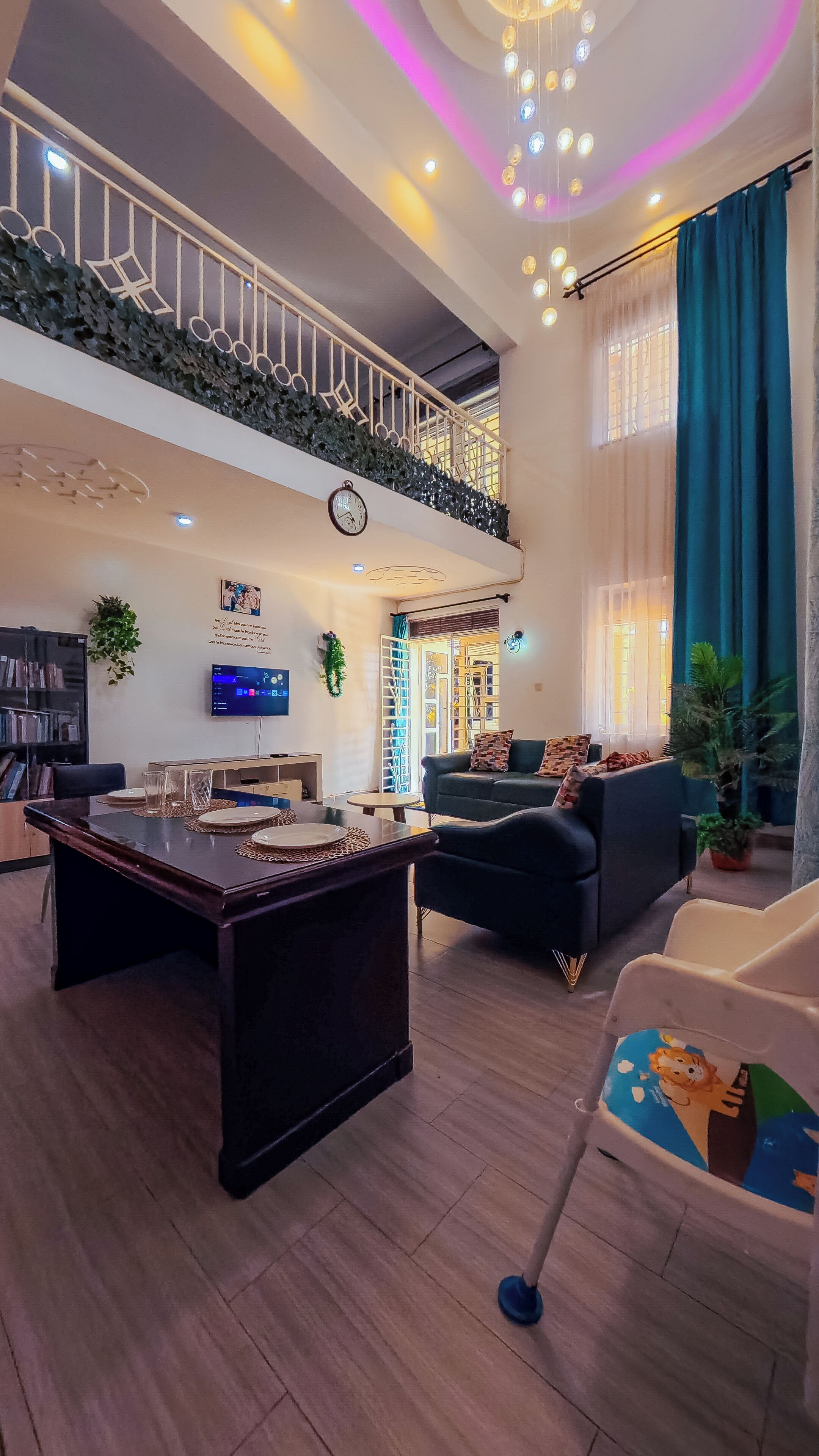 A spacious living area with a high ceiling, a dining table, a sofa, and a television mounted on the wall.