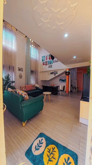 Ein Wohnzimmer mit einem grünen Sofa, einem Couchtisch und einem gemusterten Teppich. Eine Treppe führt zu einer oberen Ebene.