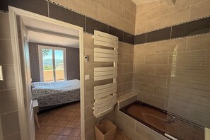 Un baño con bañera, zona de ducha y una cama visible a través de una puerta abierta.