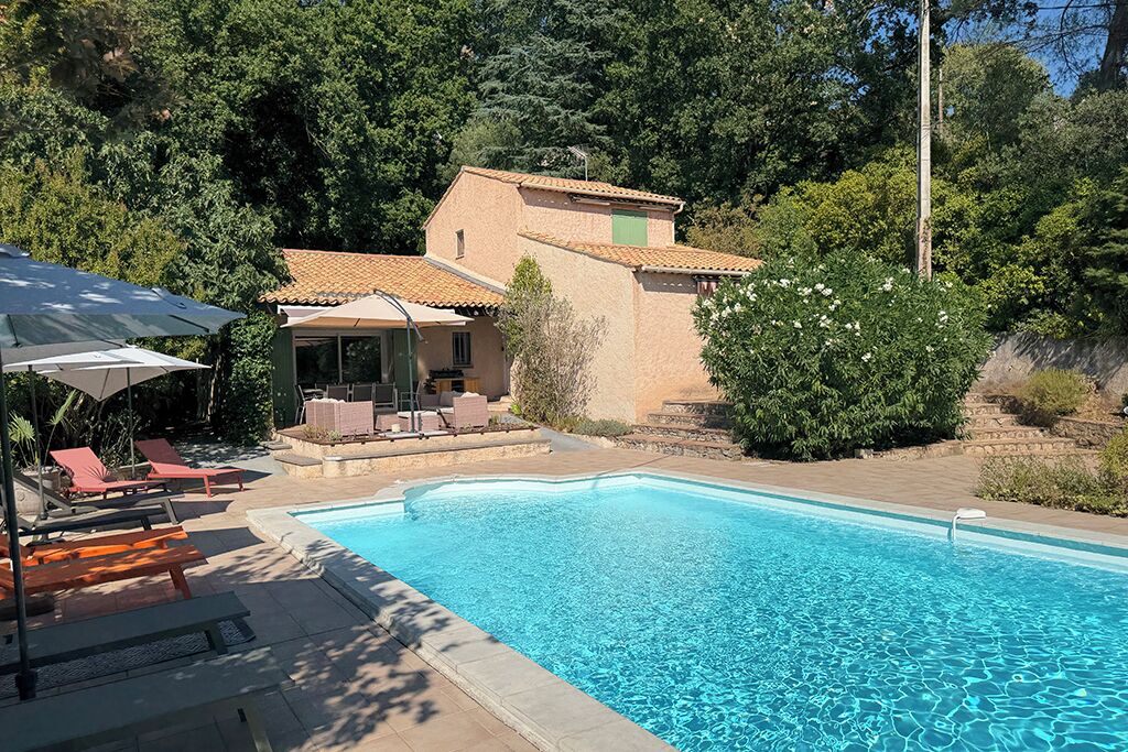 A swimming pool with lounge chairs and umbrellas, surrounded by greenery and a building.